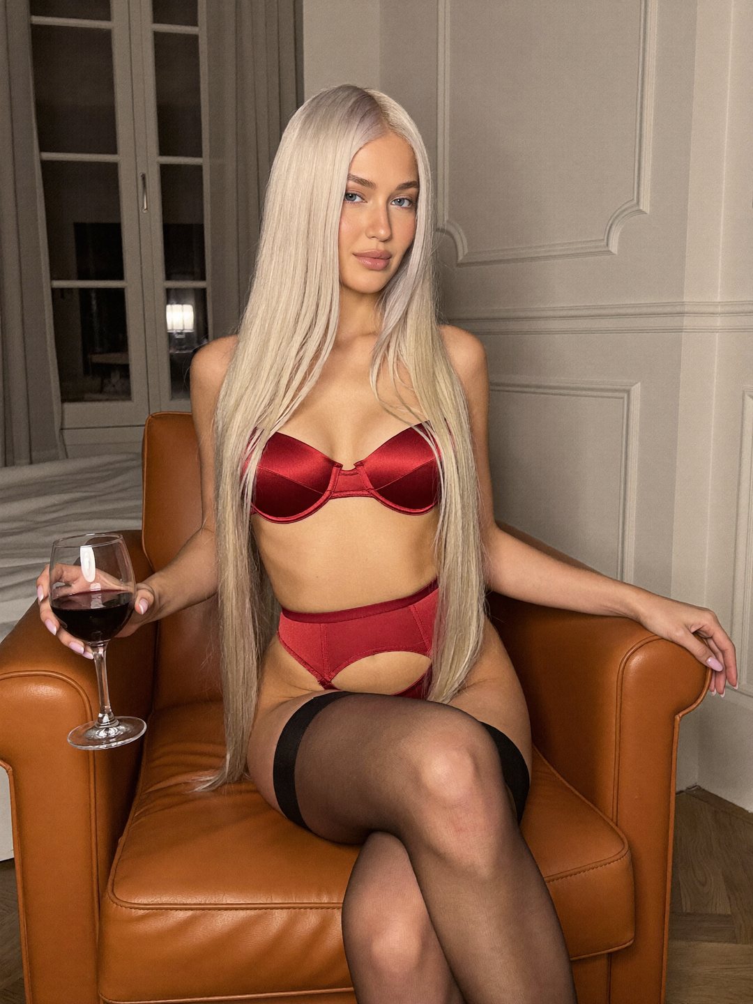 Sofiia, a blonde Ukrainian woman, sits in a cognac leather armchair with her legs crossed in a wine-red satin bra, briefs and black thigh-high stockings, holding a glass of red wine.
